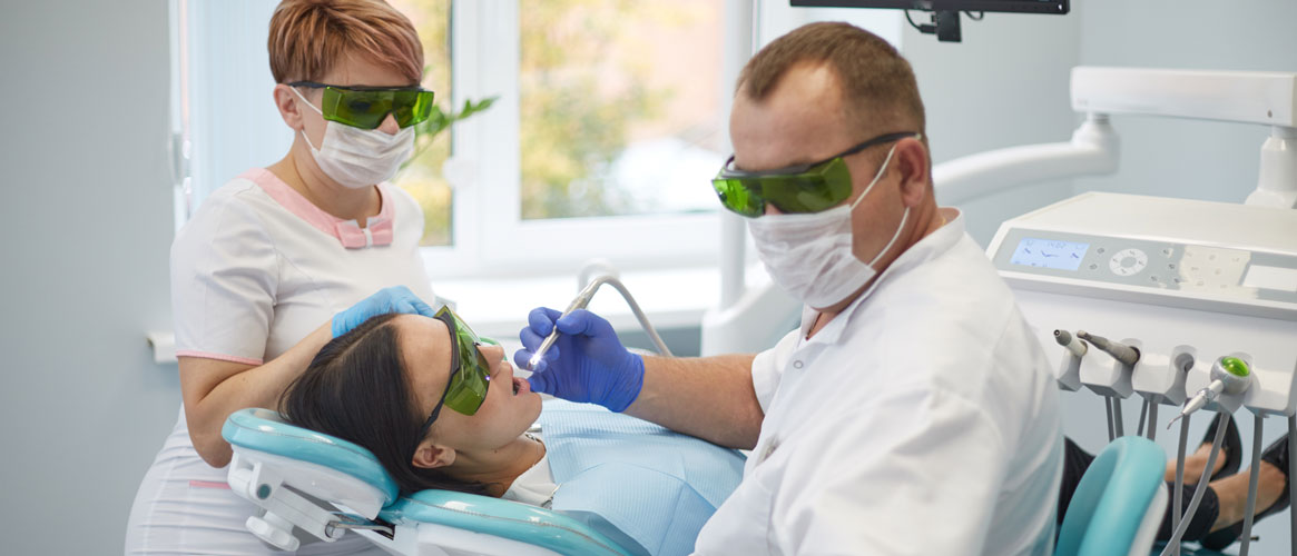 Doctor dentist treats teeth of patient while using correct ergonomic posture to prevent musculoskeletal, work-related injury. Dental hygienist looks on.