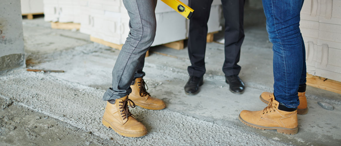 AdobeStock_137781455-two construction workers wearing jeans and brown leather work boots standing with man in suit on concrete floor