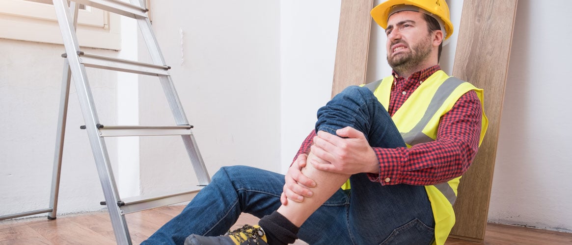 Construction worker holding shin on floor next to ladder after painful accident