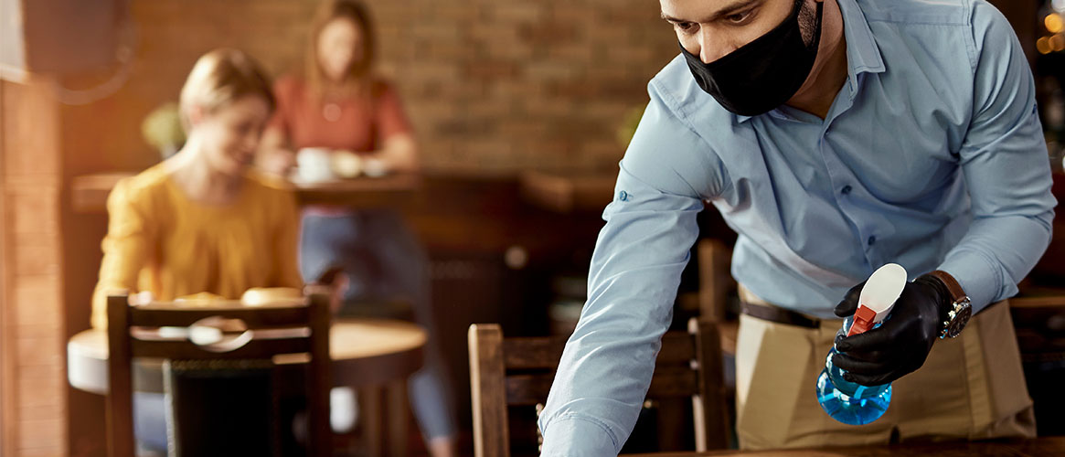 Restaurant worker wearing mask cleaning table, as customers dine in the background
